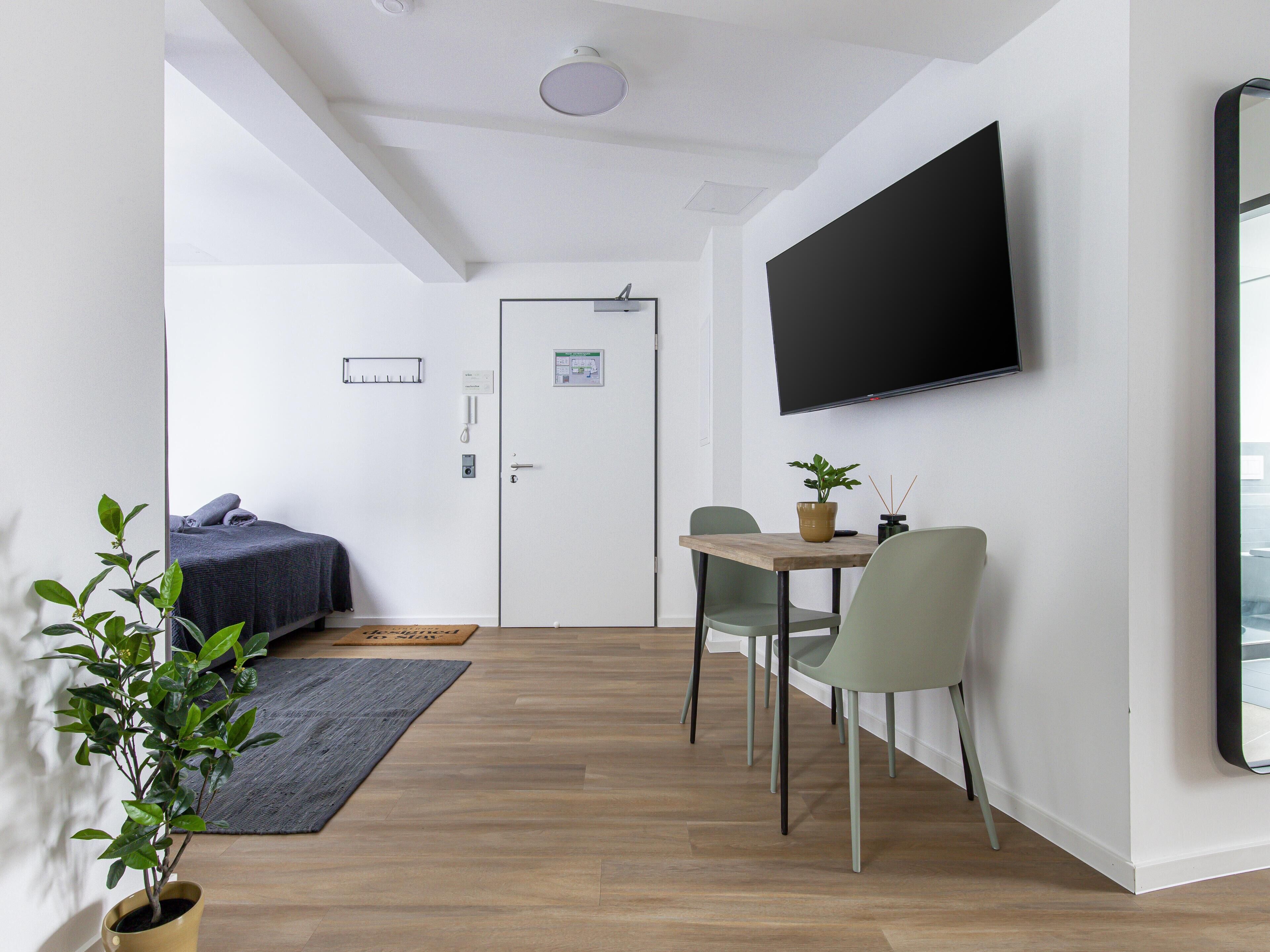 Una habitación moderna y bien iluminada con una cama, una mesa de comedor con sillas, una planta en maceta y un televisor de pantalla plana montado en la pared.