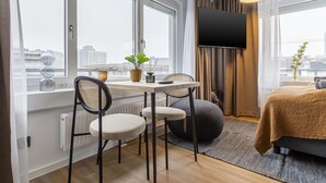 A modern hotel room with a large window, a flat-screen TV, a dining table with chairs, a sofa, and a potted plant. - Limehome Essen Kettwiger Str. (Essen)