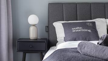 Ein Schlafzimmer mit grauem Kopfteil, einem Nachttisch mit Lampe, einem Bett mit grauer Bettwäsche und einem gerahmten Bild an der Wand.