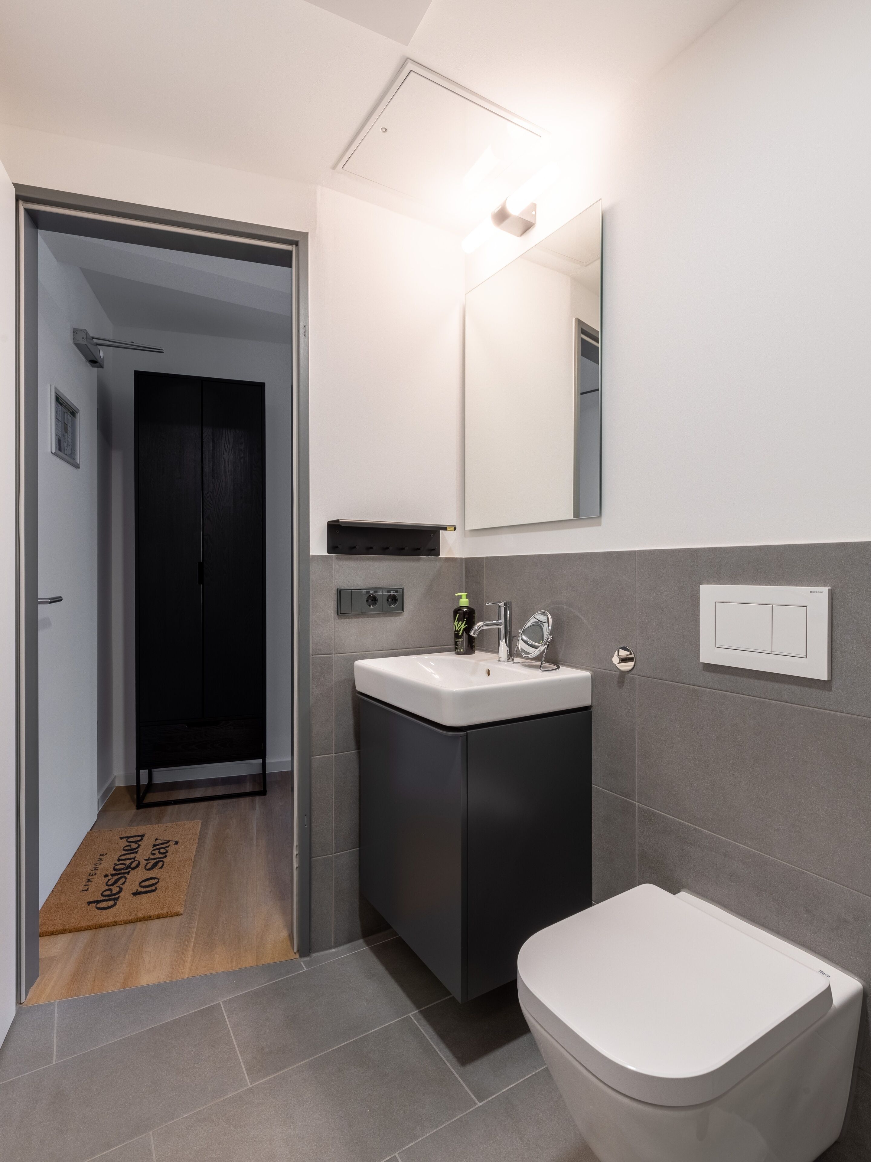A modern bathroom with a white sink, a toilet, and a mirror. There is a black cabinet below the sink and a door leading to another room.