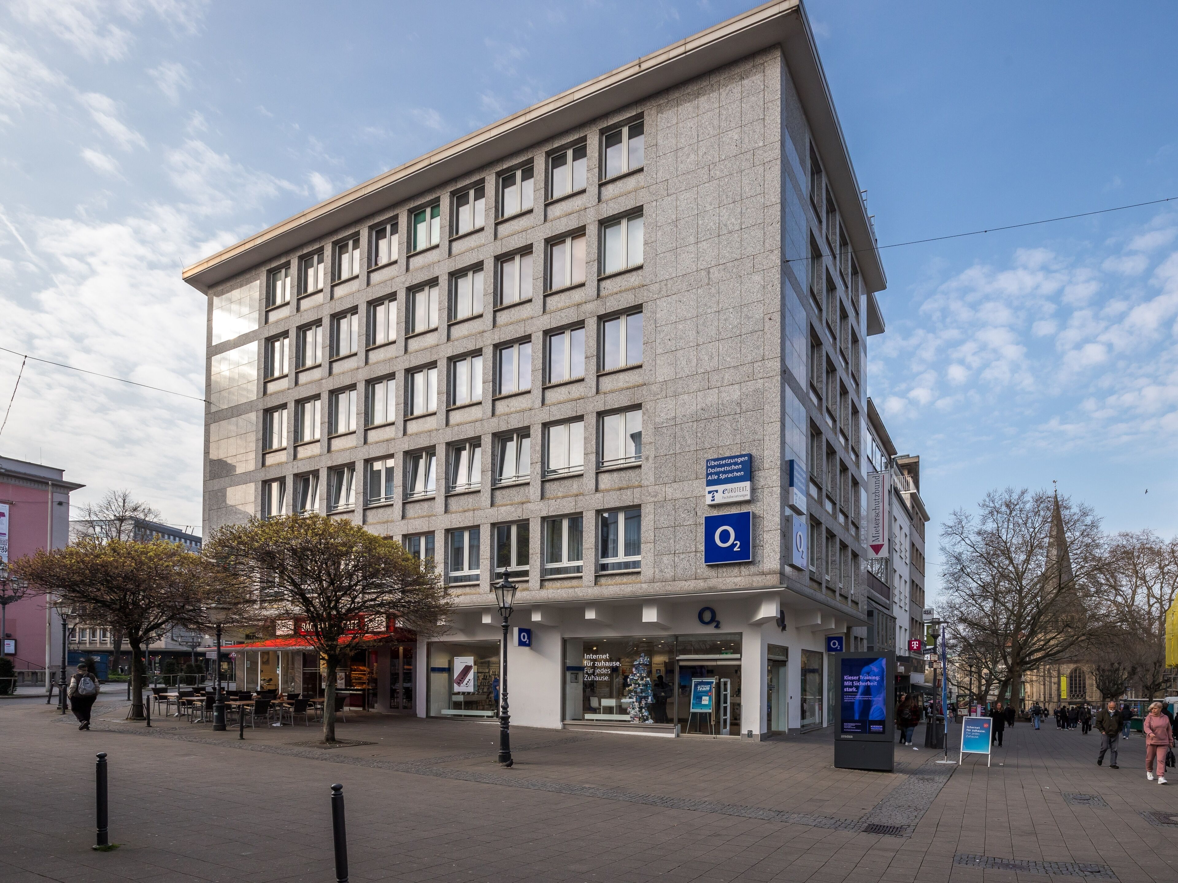 Un edificio moderno con una fachada de ventanas enrejada, un letrero que indica un hotel Q+Q, y una zona peatonal con árboles y cielo despejado.