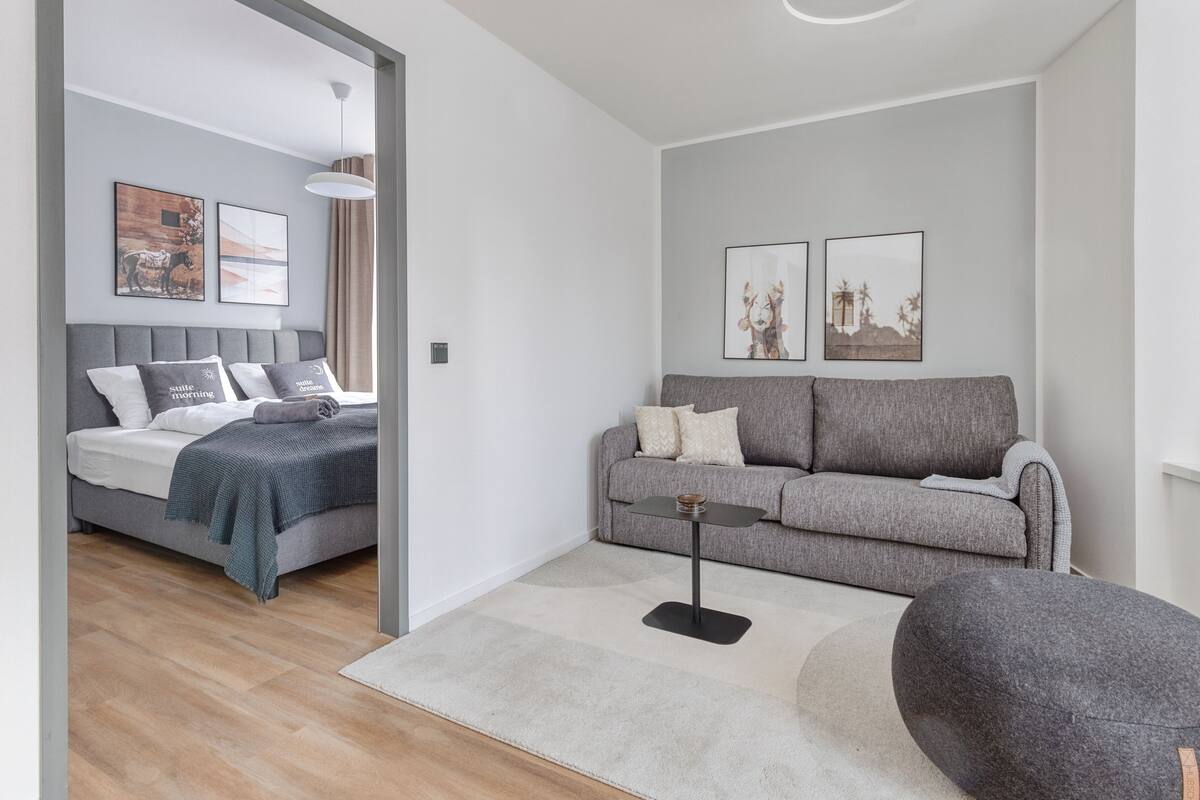 Una sala moderna con un sofá gris, un sillón gris y una cama con cabecero gris. Hay dos cuadros enmarcados en la pared, una mesita con una lámpara y un otoman gris.