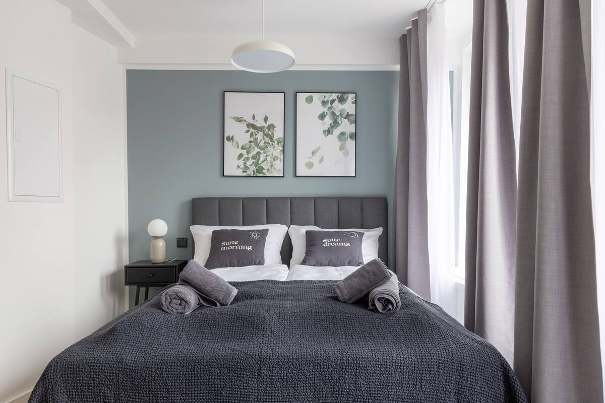 Una cama bien hecha con una manta gris y dos almohadas, una mesita de noche con una lámpara, y dos láminas botánicas enmarcadas en la pared.