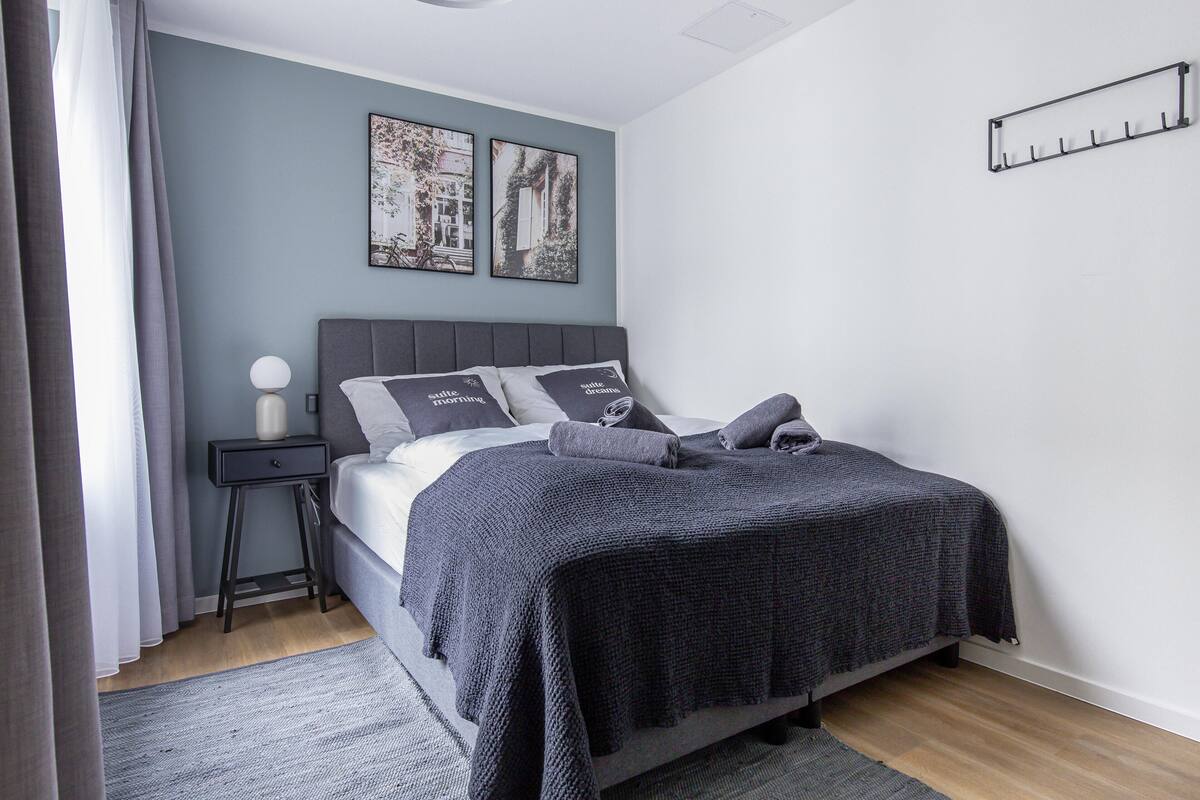Un dormitorio moderno con cabecera gris, colcha gris y dos almohadas a juego. Hay una mesita de noche con una lámpara y un estante montado en la pared.