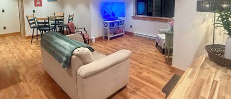 A living room with a beige sofa, wooden flooring, a TV, and a window.