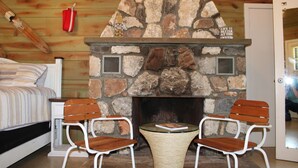 A cozy room with a stone fireplace, two wooden chairs, a small table, and a bed with striped bedding.