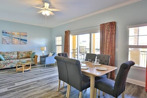 A dining area with a table set for four, surrounded by chairs, in a room with a ceiling fan, a sofa, and a view through large windows.