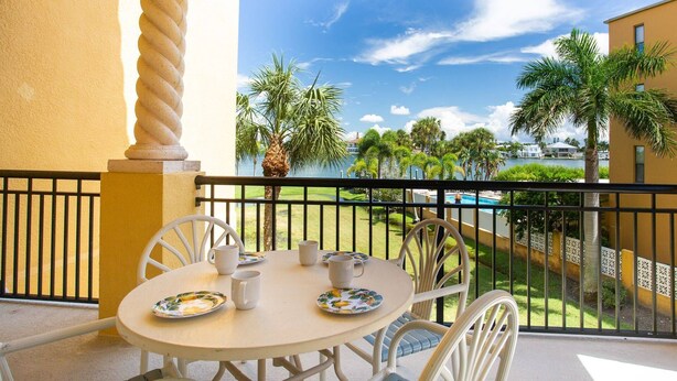 A round table with chairs and a small table setting on a balcony with a view of a pool and palm trees. - Two Private Balconies! Heated Waterfront Pool & Hot Tub, Fish From the Dock, Just Steps to the Beach (Treasure Island)