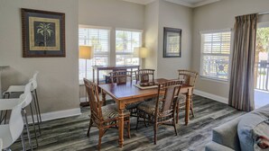 A dining area with a wooden table and wicker chairs, a white sofa, and two windows with blinds. - Two Private Balconies! Heated Waterfront Pool & Hot Tub, Fish From the Dock, Just Steps to the Beach (Treasure Island)