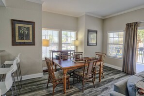 A dining area with a wooden table and wicker chairs, a white sofa, and two windows with blinds.
