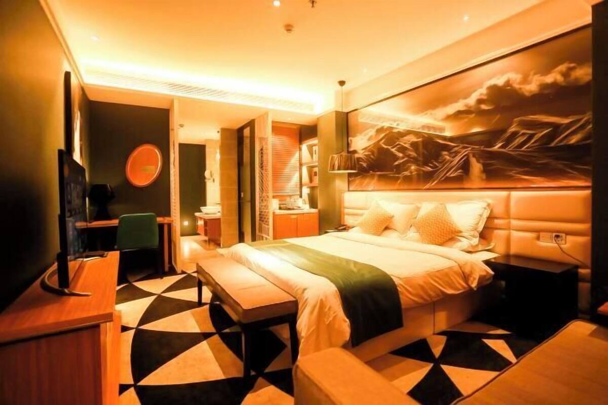 Quarto de hotel com cama, escrivaninha, cadeira, televisão e um quadro na parede.
