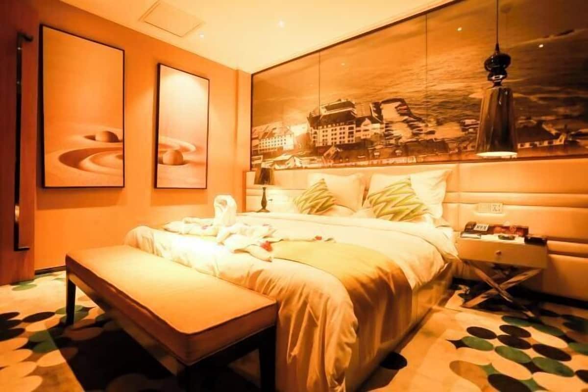 Quarto de hotel com uma cama grande, um banquinho, uma mesa de cabeceira e uma obra de arte na parede.
