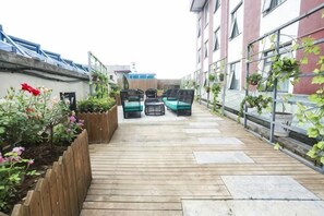 Una terraza de madera con muebles de exterior, un cantero con flores y un edificio con ventanas y balcones.