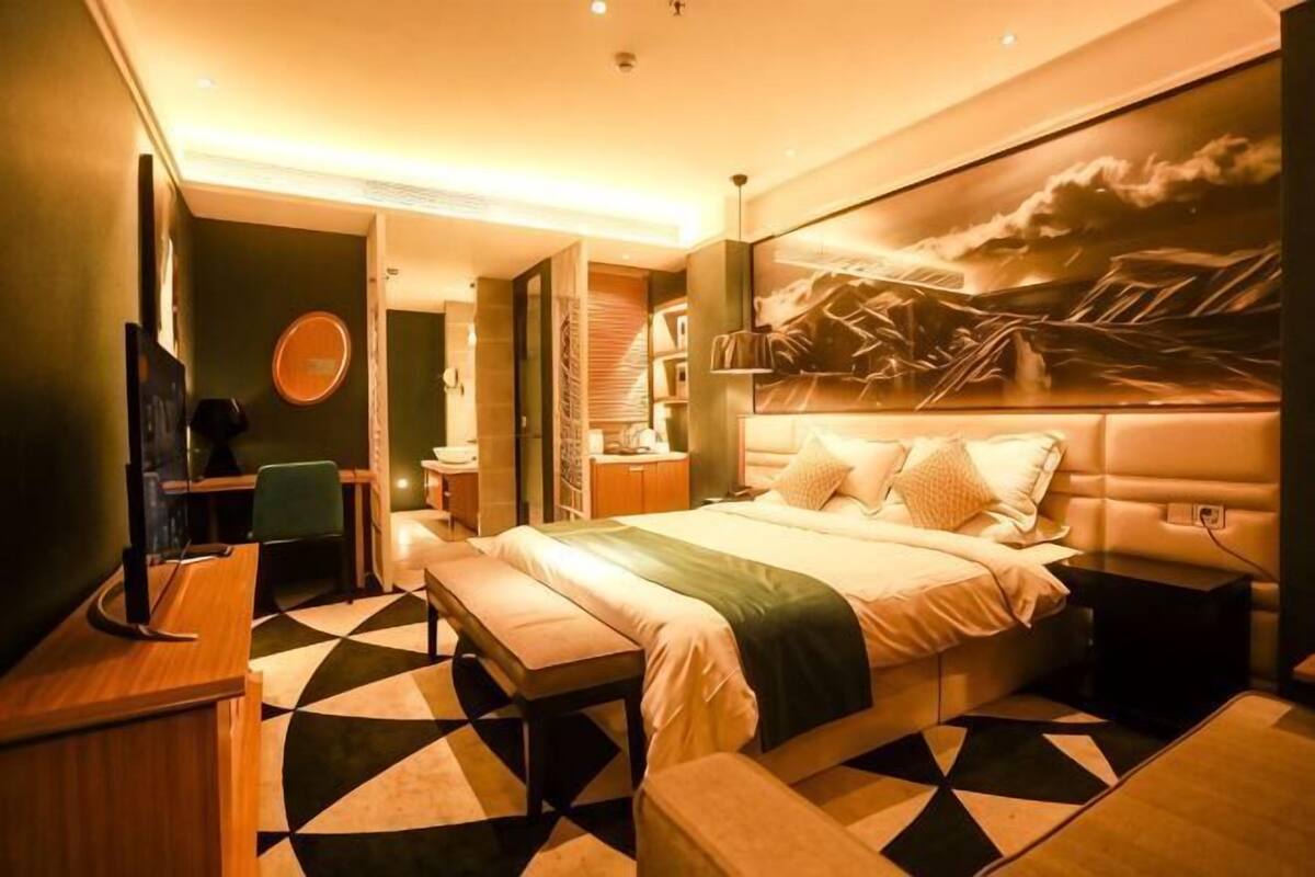 Quarto de hotel com cama, escrivaninha, cadeira, televisão e um quadro na parede.