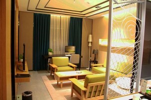 Habitación de hotel con escritorio, televisor, cama con cabecera blanca y zona de estar con sillas y una mesa.