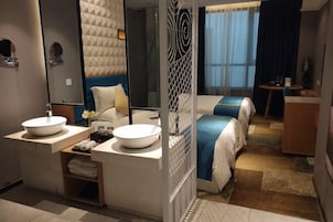 Una habitación de hotel moderna con dos camas, un espejo grande, un lavamanos redondo y una mesita pequeña.
