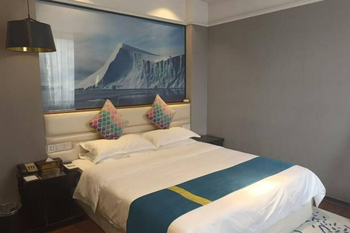 Quarto de hotel com uma cama grande, uma mesa de cabeceira com telefone e uma pintura de uma paisagem de montanha na parede.