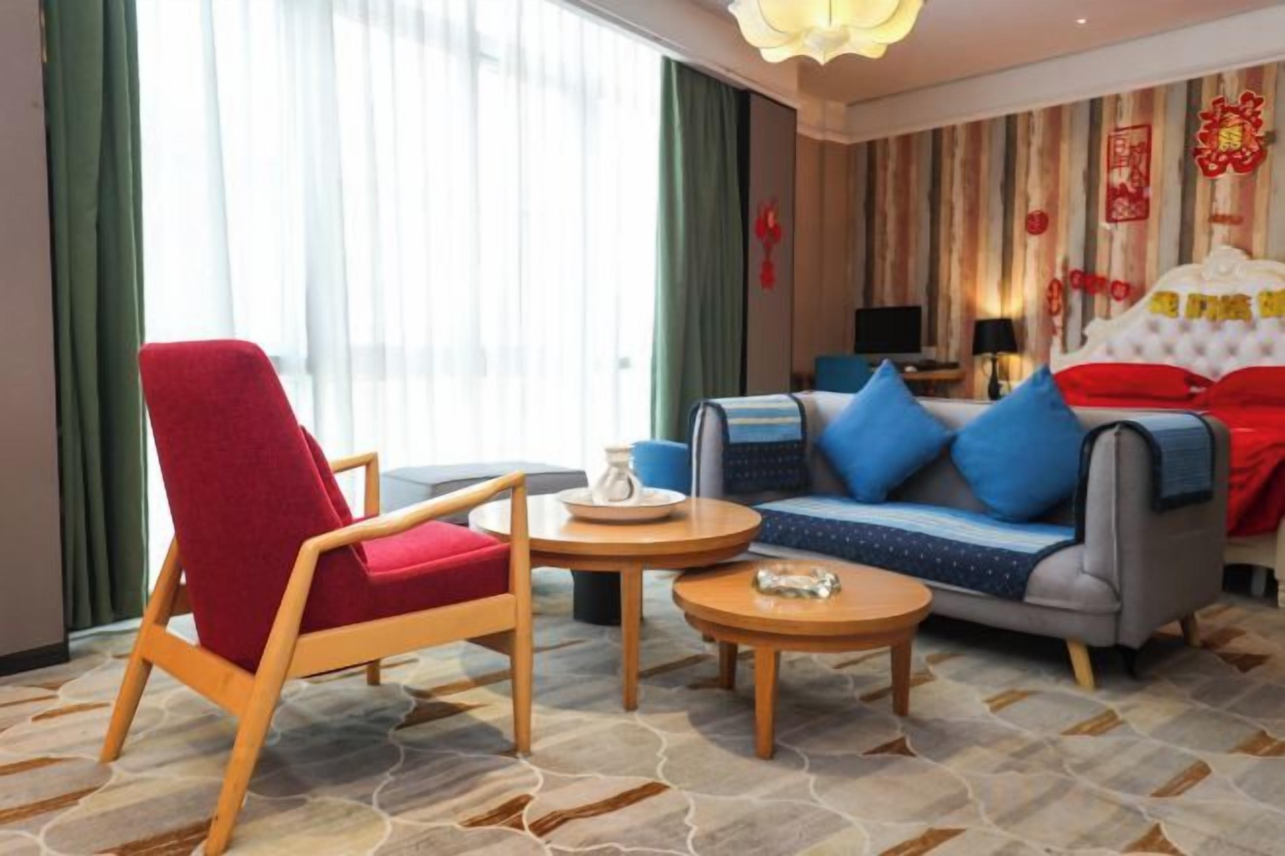 Ein Hotelzimmer mit einem roten Sessel, einer blauen Sitzsofa mit Kissen, einem kleinen runden Tisch und einem Bett mit roter Bettwäsche.