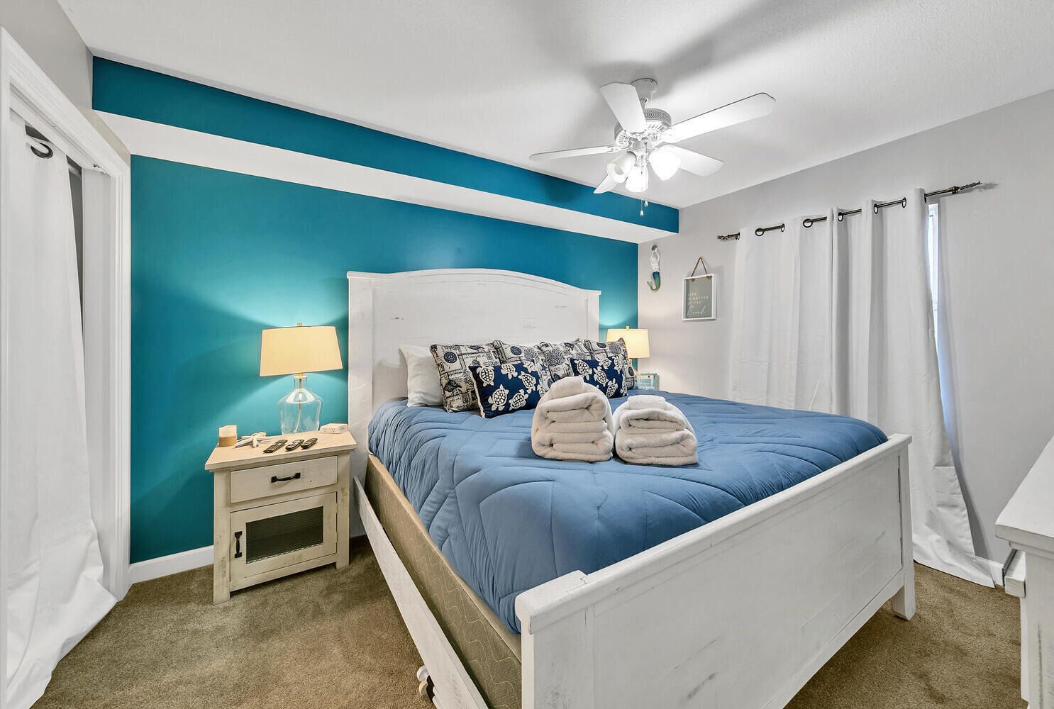 A bedroom with a white bed, teal walls, a ceiling fan, and a nightstand with a lamp.