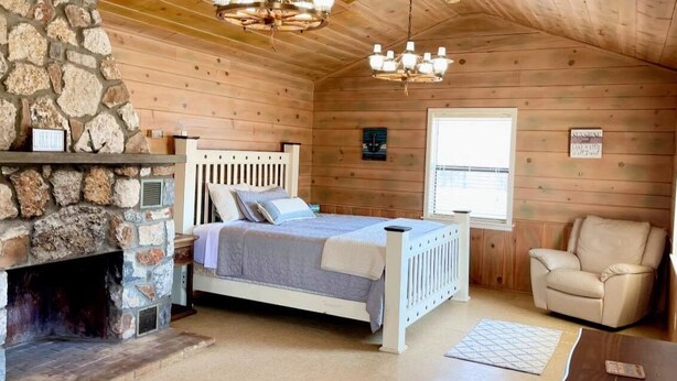 A cozy bedroom with a stone fireplace, a bed with a headboard, a window, and a chair. - Grand lake Cabin 3 @ Stony Point (Jay)