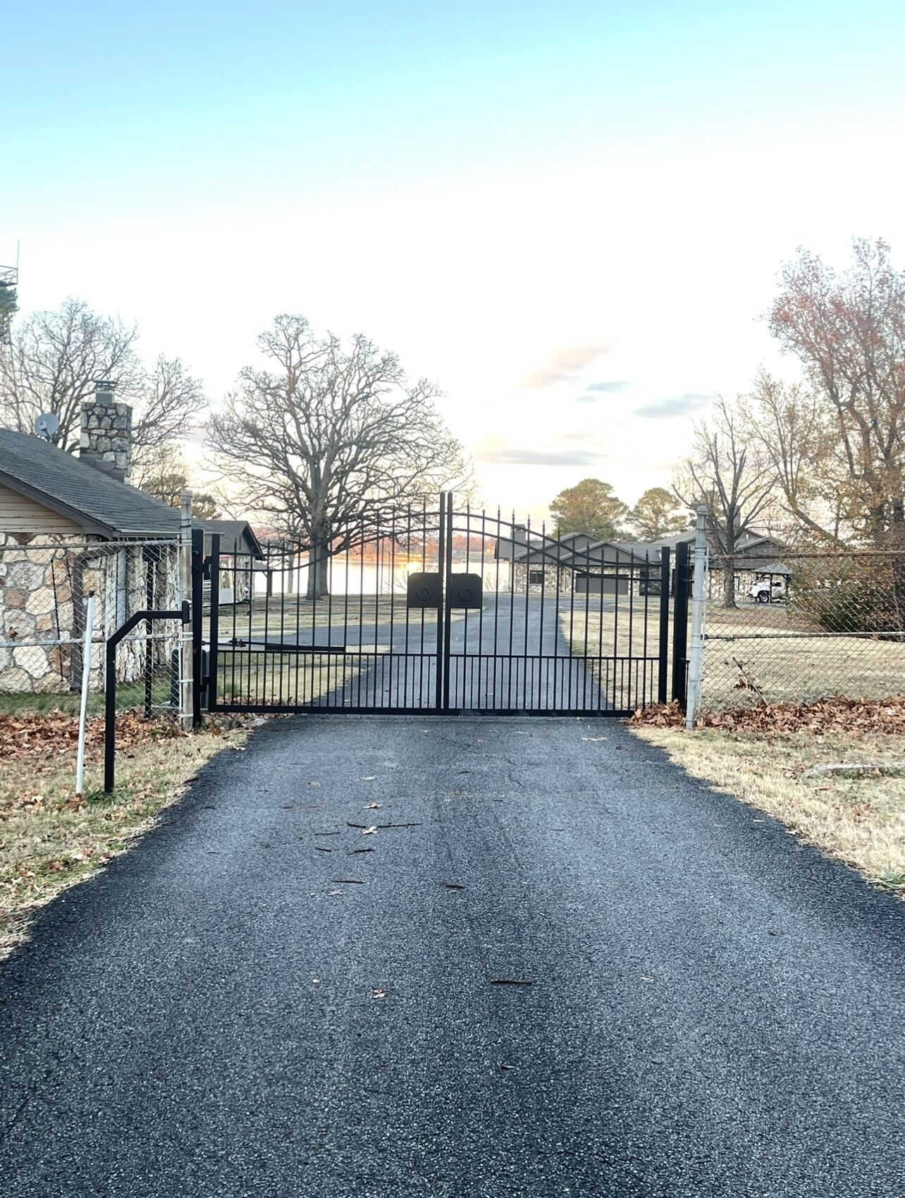 A gated driveway leading to a property with a stone building on the left and a fenced area with trees.