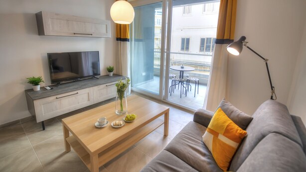 A modern living room with a sofa, coffee table, TV, and a balcony with a view of buildings. - Bright and modern apartment near seafront BFRA1-2 (Gzira)