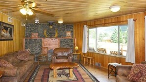 Ein gemütliches Blockhauss-Interieur mit Kamin aus Stein, Ledersofas und einem großen Fenster mit Blick ins Freie.
