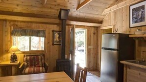 A wooden cabin interior with a dining table, chairs, a refrigerator, a stove, and a window with curtains. - Comfy cabin is situated on the Silver Gate property which overlooks 10,000ft tall mountain peaks surrounding the town. (Silver Gate)