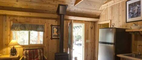 A wooden cabin interior with a dining table, chairs, a refrigerator, a stove, and a window with curtains.