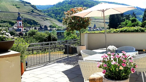 Una terrazza con mobili in vimini, una pianta in vaso con fiori rosa e vista su una chiesa e vigneti sullo sfondo.