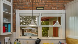 A kitchen with a brick wall, white cabinets, a window with a view of a pool, and a bowl of lemons and chips on the counter.