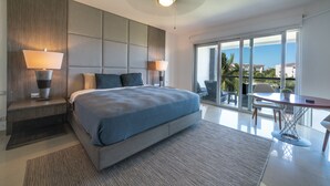 Une chambre moderne avec un grand lit, deux lampes de chevet, une vue sur l’extérieur grâce à de grandes fenêtres, et un coin repas avec une table et des chaises.