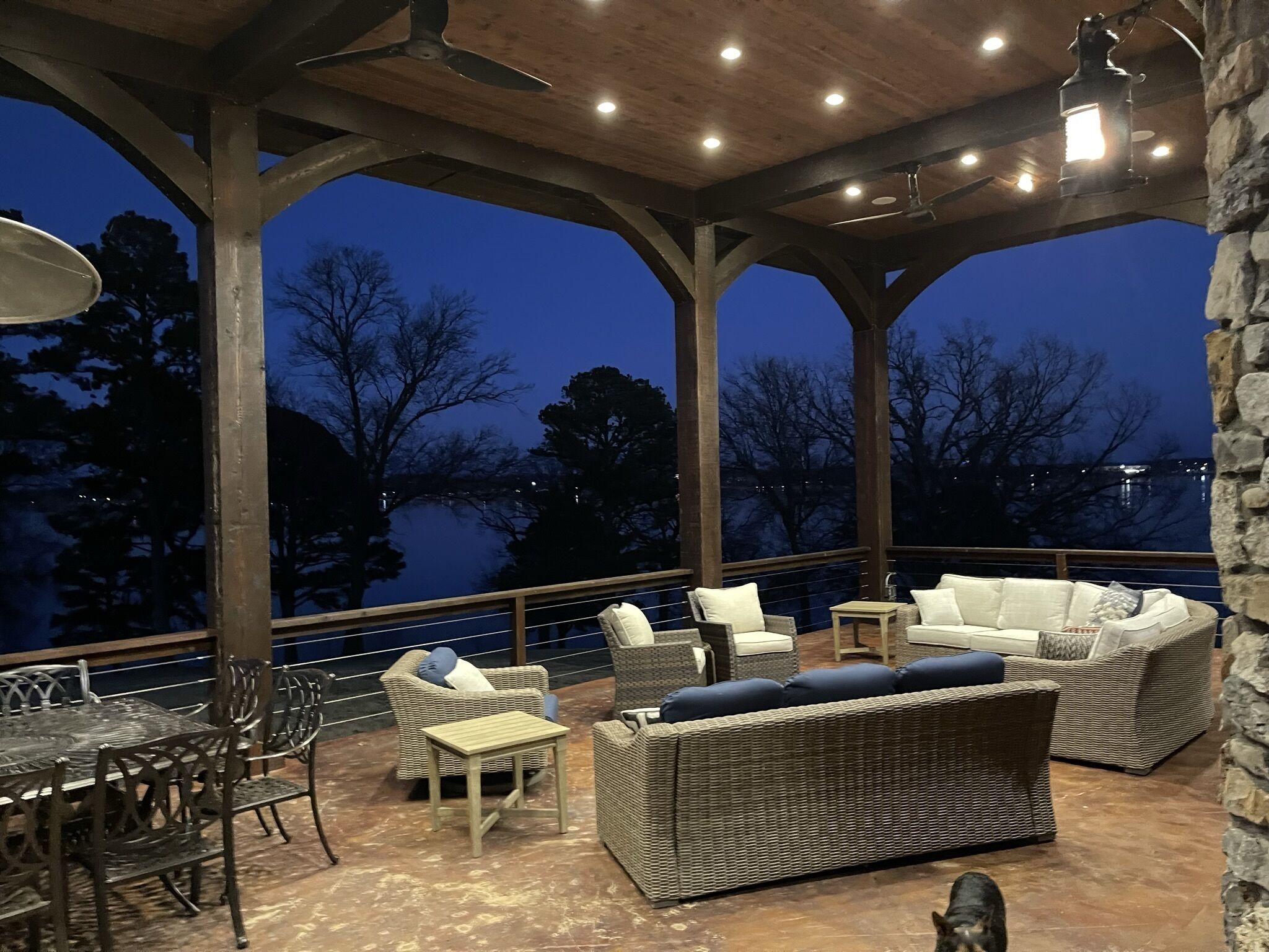 An outdoor patio with wicker furniture, a dining area, and a view of trees and a body of water at dusk.