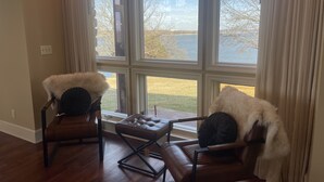 Una habitación con dos sillones de cuero y un pequeño reposapiés, un ventanal con vistas al agua y árboles, y suelo de madera.