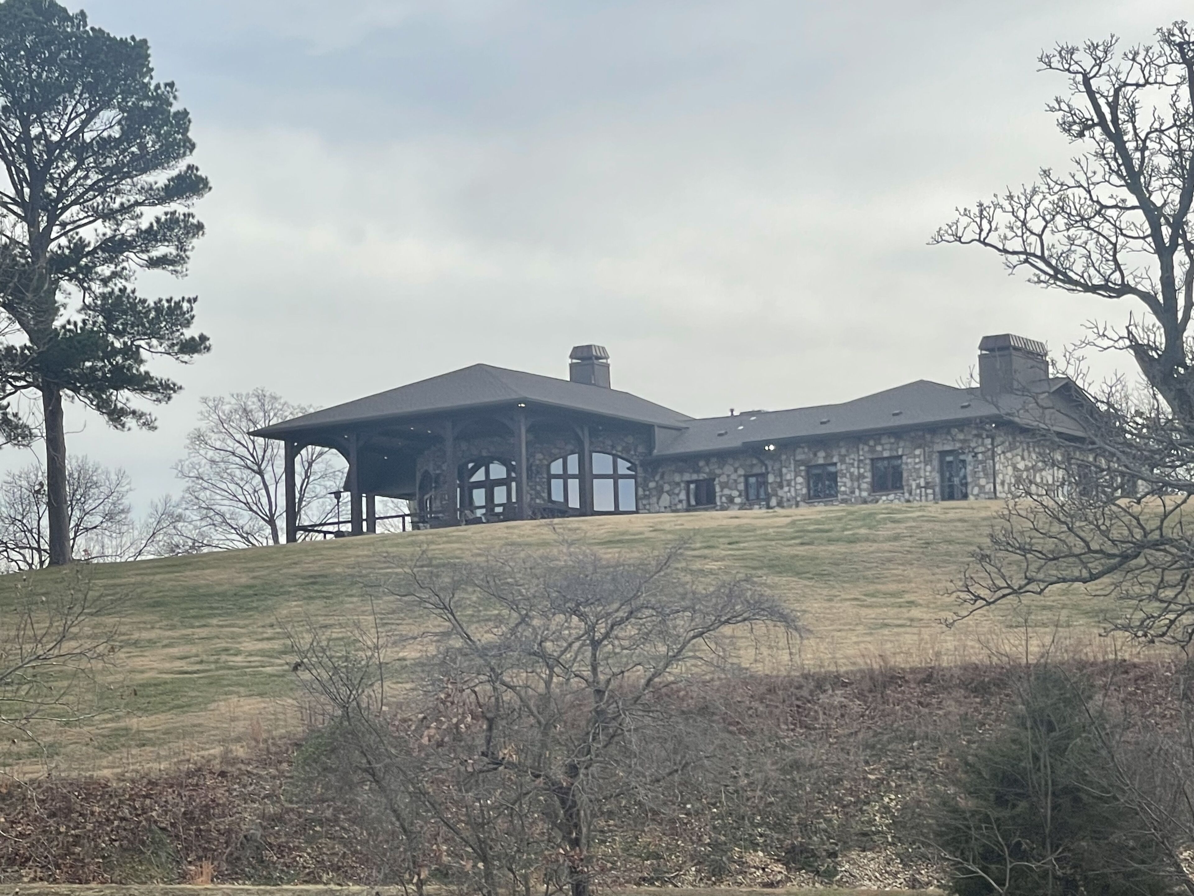 Une grande maison en pierre, dotée d’une imposante cheminée en pierre, d’un porche couvert et offrant une vue sur une colline verdoyante.