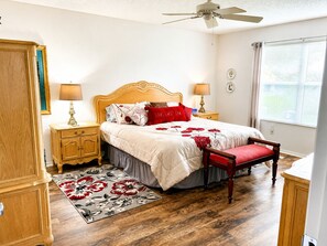 A bedroom with a wooden bed, bedside tables, a nightstand, a red chair, a ceiling fan, and a window with blinds. - CLEAN, CLEAN, CLEAN; Walk to pools; Golf Cart Included; NO Xtra guest fees !!! (The Villages)