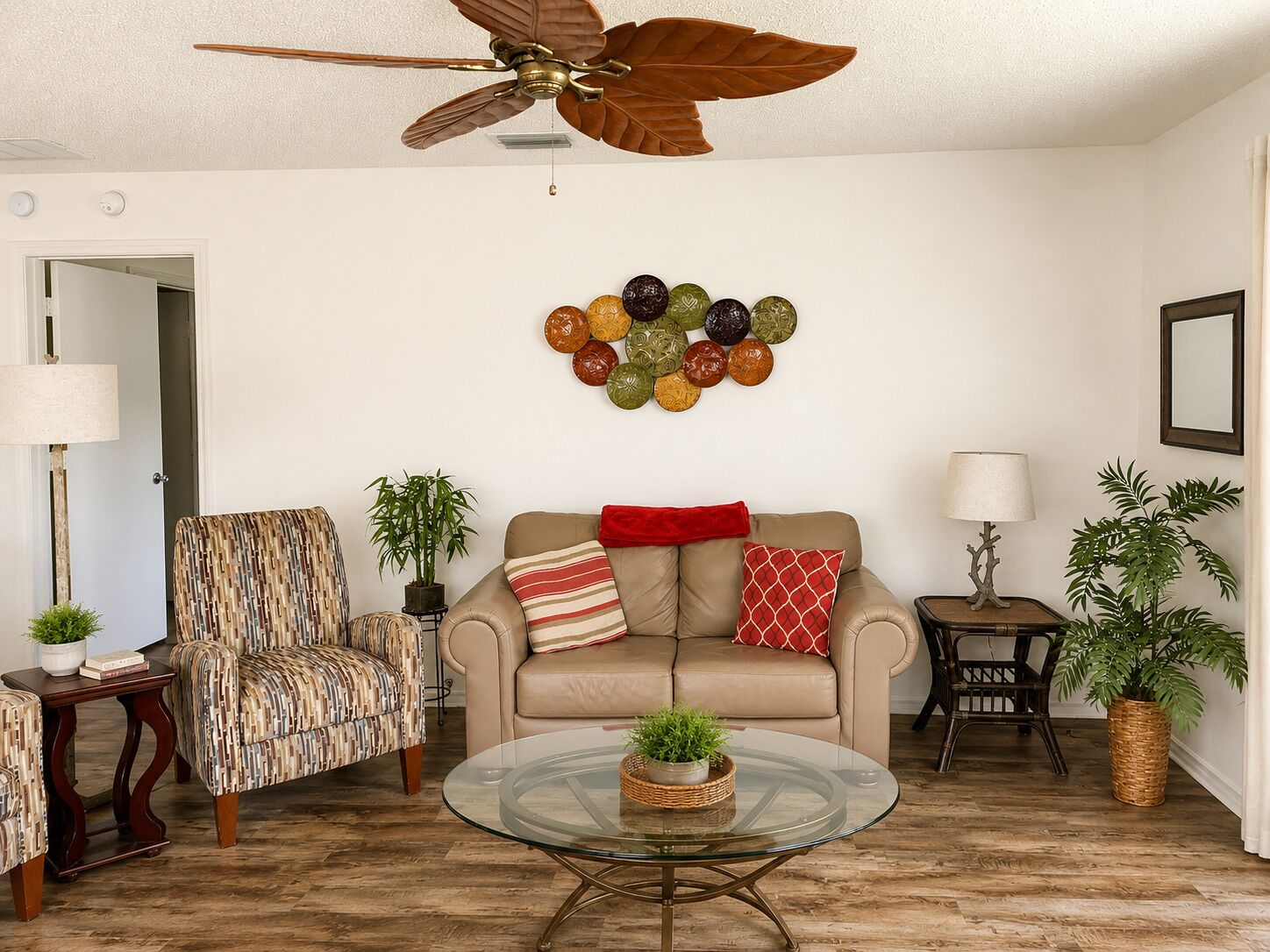 A living room with a ceiling fan, a sofa, a patterned armchair, a glass coffee table, and decorative wall art.