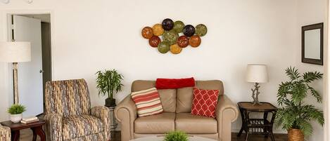 A living room with a ceiling fan, a sofa, a patterned armchair, a glass coffee table, and decorative wall art.
