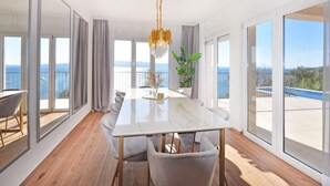 A modern dining room with a large table, comfortable seating, and a view of the sea through large windows. - Villa Iva With sea View and Pool (Omis)