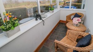 A corner room with a large window overlooking a rural landscape, wicker furniture, and a small table with flowers. - Twincote (Wooler)
