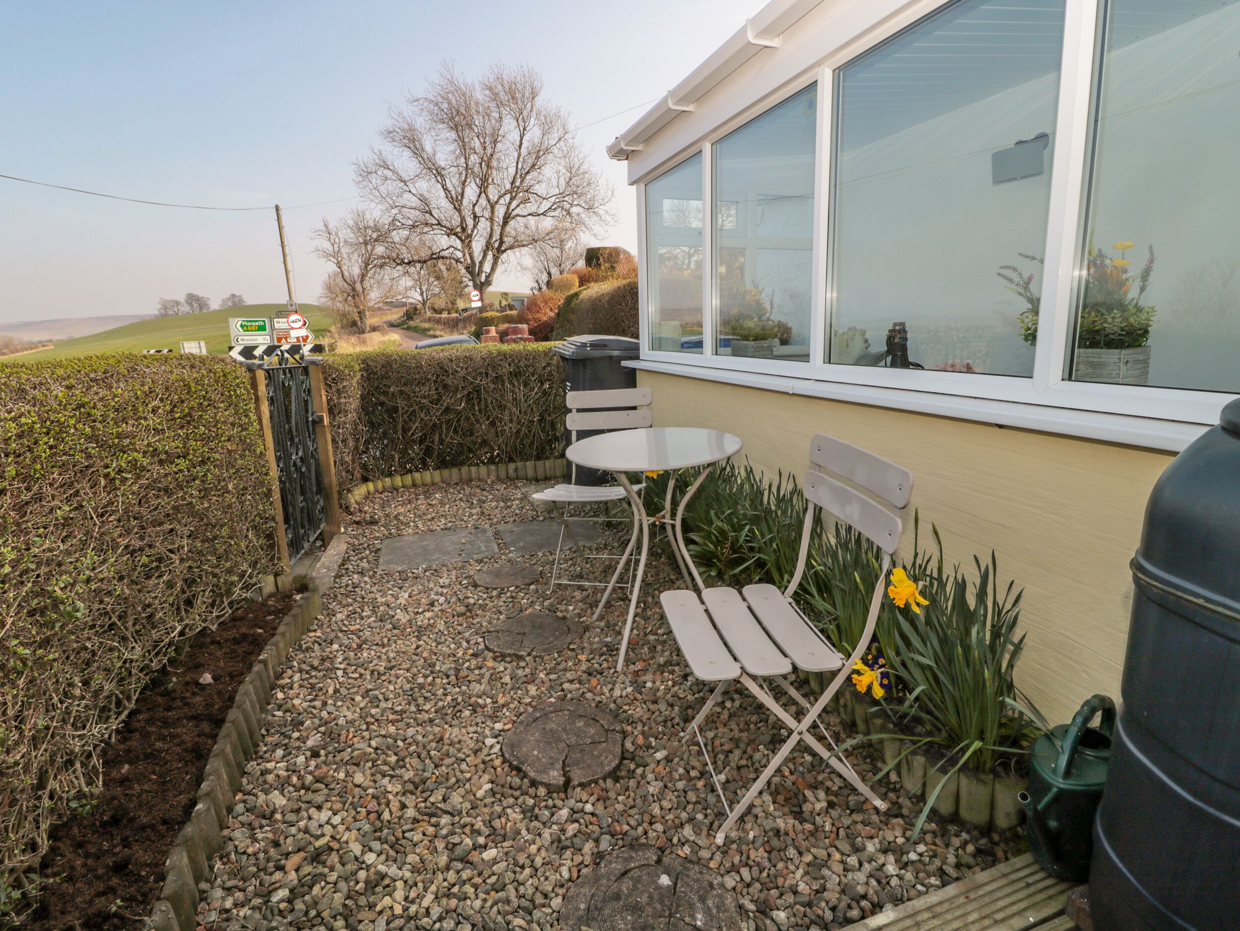An outdoor patio with a table and chairs, a large window, and a garden area with plants and a hedge.