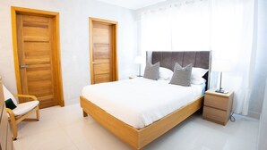 A bedroom with a wooden bed, two doors, a chair, a bedside table with a lamp, and a window with curtains. - Beautiful 2BR City Center - Renty (Santo Domingo)