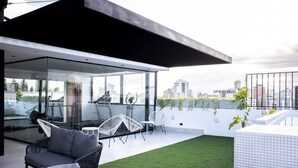 A modern rooftop terrace with glass walls, outdoor seating, and a view of the city skyline. - Beautiful 2BR City Center - Renty (Santo Domingo)