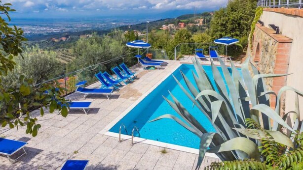 Zona de piscina con sillas reclinables azules y sombrillas, rodeada de vegetación y con vistas a un paisaje pintoresco.