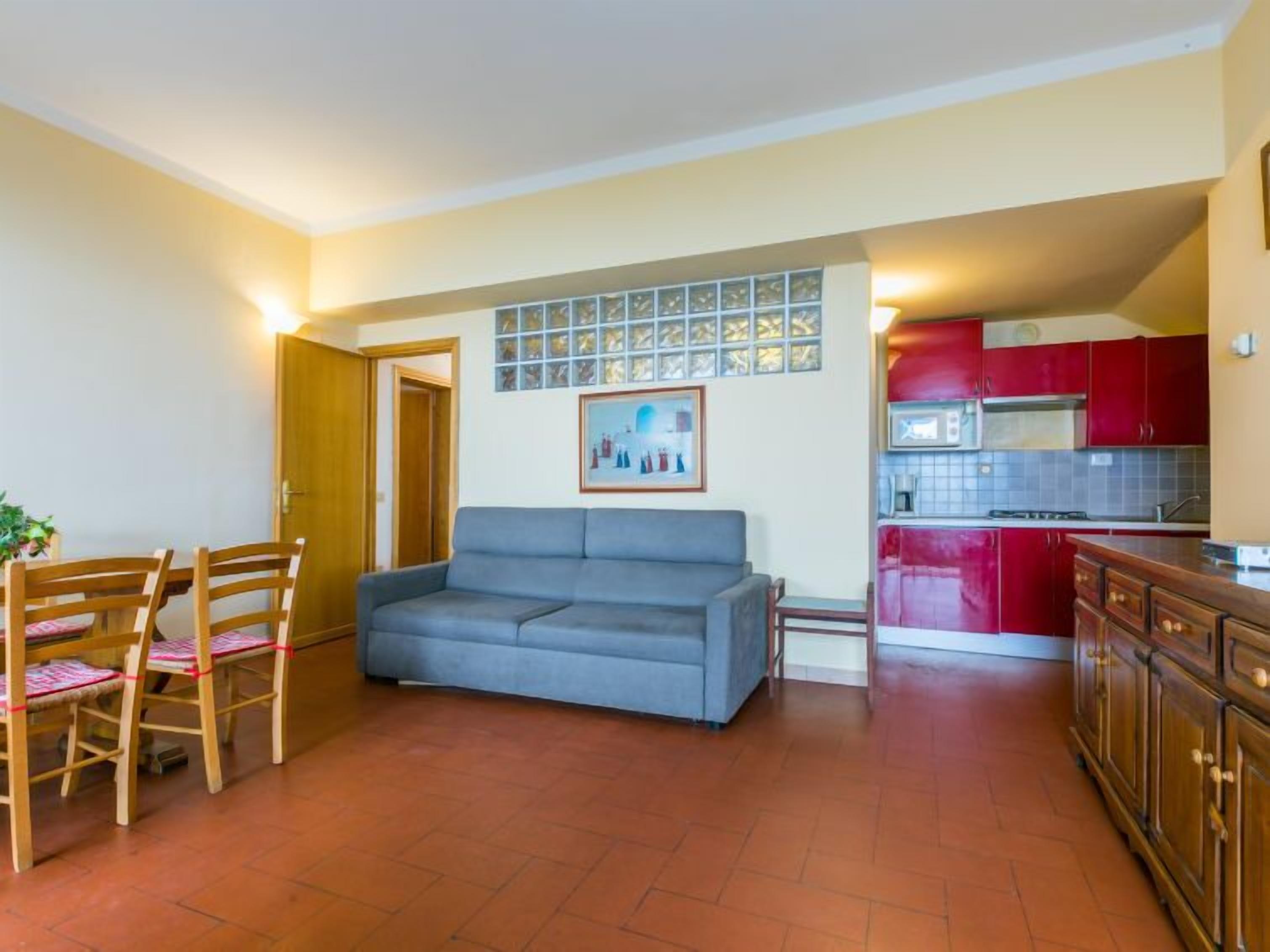 Una sala de estar con un sofá gris, sillas de madera, una mesa de comedor y una cocina con armarios rojos.