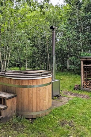 A wooden hot tub with a metal chimney, surrounded by a grassy area and trees. - Riverside Cottage by Interhome (Drumnadrochit)