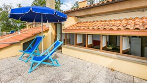 Un hotel con tetto in tegole, una zona relax all'aperto coperta, con sedie blu e un ombrellone, e una finestra con tende.