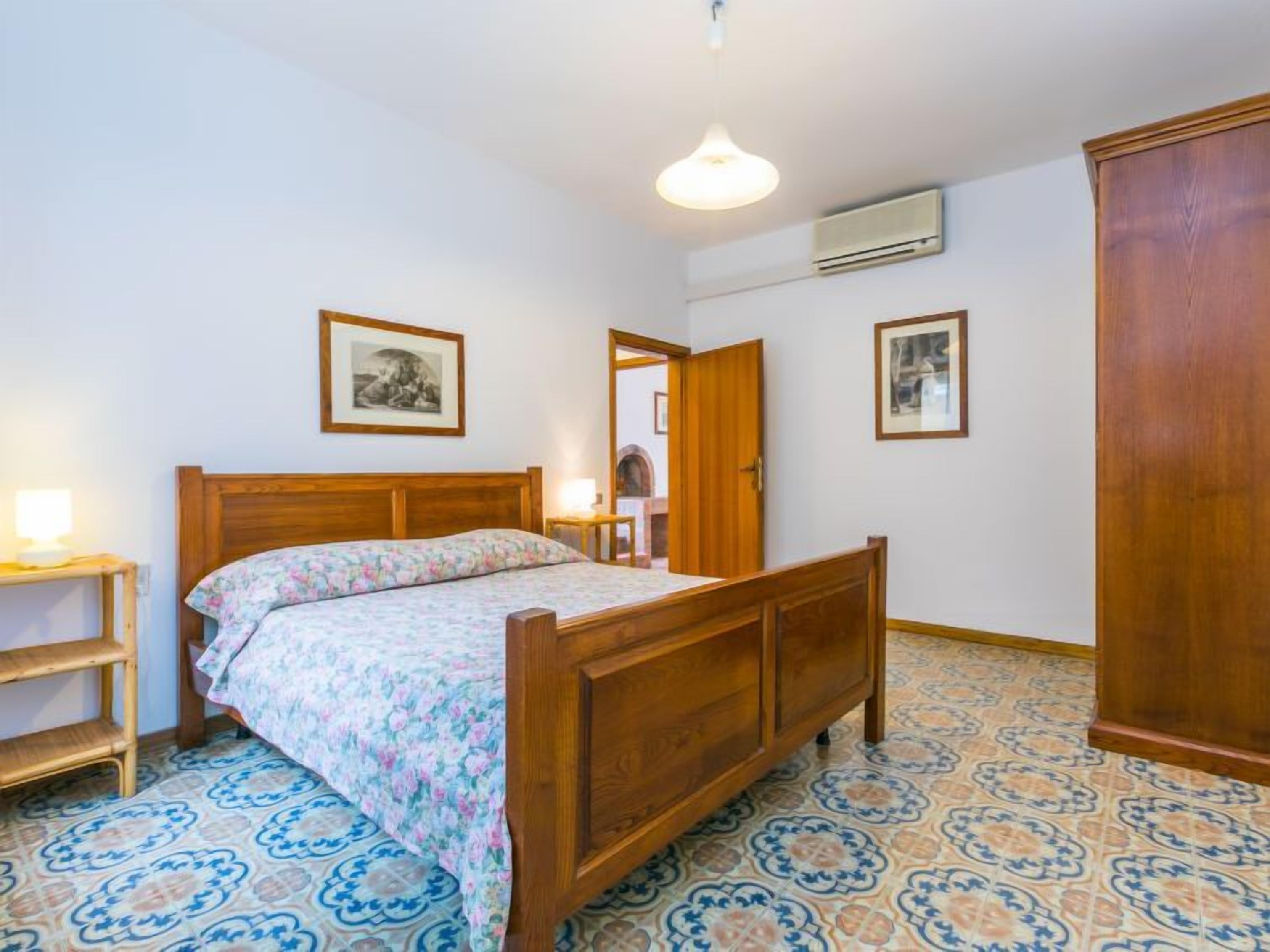 A bedroom with a wooden bed, a nightstand, a wardrobe, and patterned floor tiles.