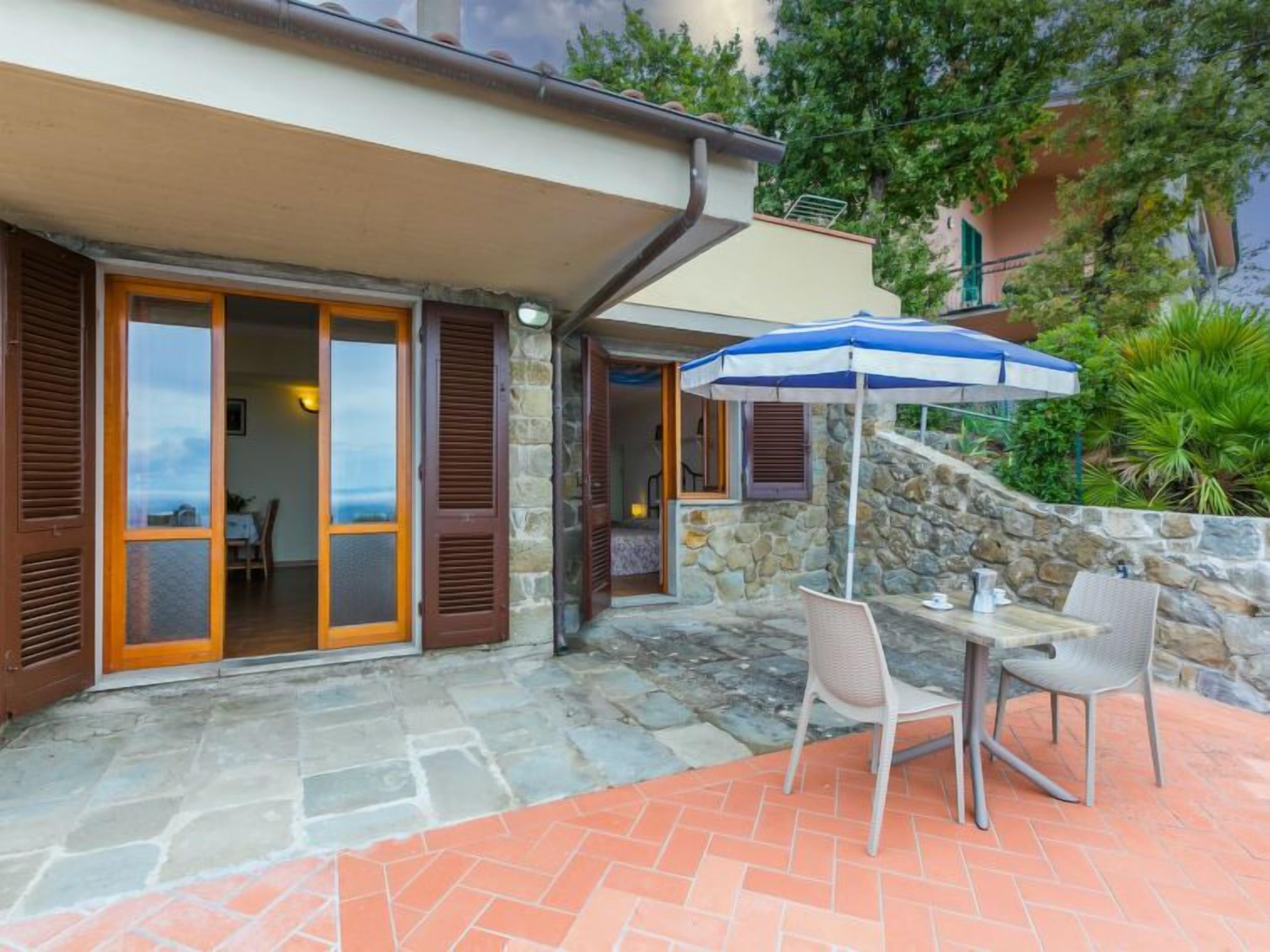 Une terrasse dallée de pierres, avec une table et des chaises, un parasol bleu et blanc, et une vue sur la mer.