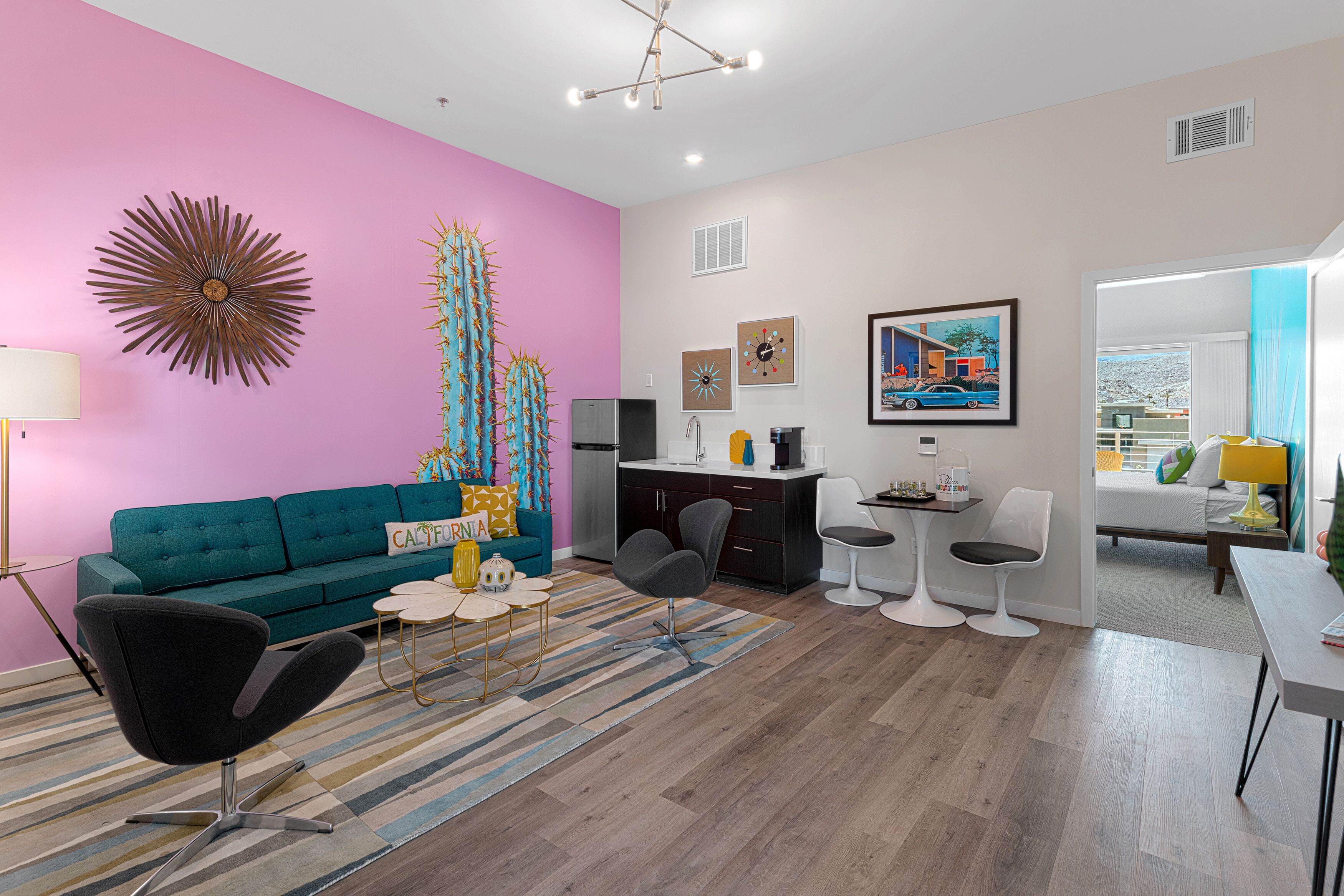 A modern living room with a teal sofa, a black armchair, and a coffee table. There is a kitchen area with a refrigerator, stove, and sink. A dining area with white chairs and a table is visible.